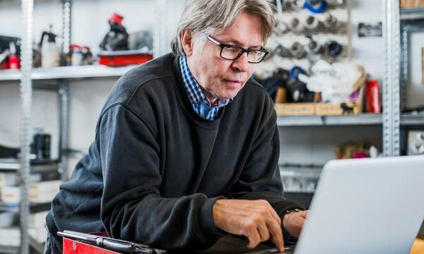 An older man with medium-length gray hair staring pensively at a silver laptop screen. He’s wearing a black crew neck sweater over a blue and white checkered shirt. The background is a blurred workshop with wall mounts and shelving units.